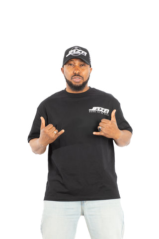 „Schwarzes JAZA Born To Be Rich Unisex T-Shirt, getragen von einem Model, frontal aufgenommen mit Handgesten vor weißem Hintergrund.“